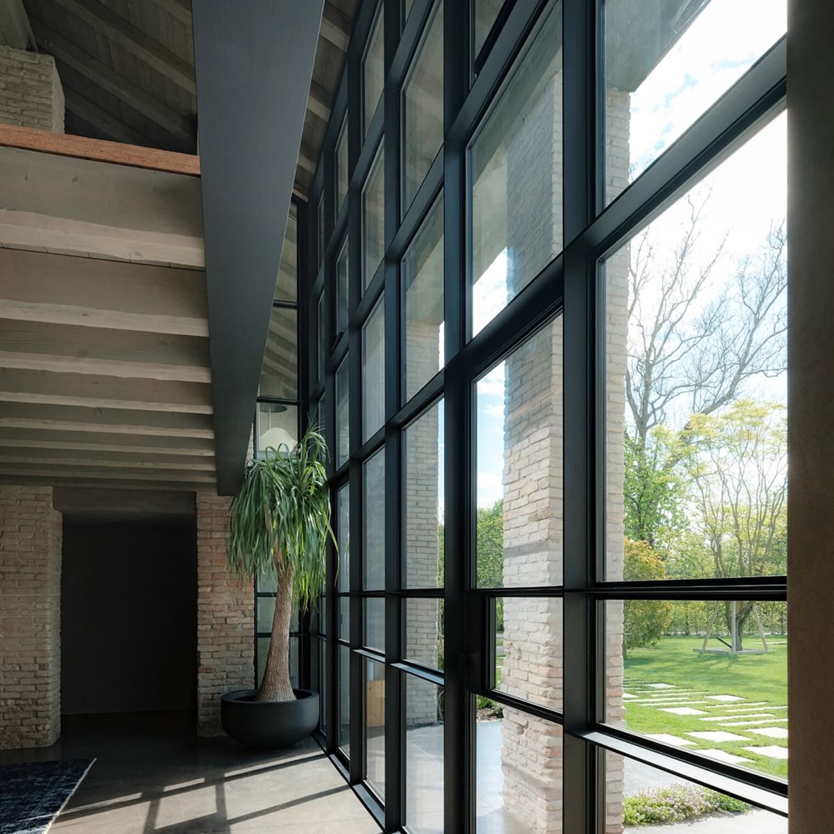 Double-height black steel window wall along a brick corridor overlooking a green lawn